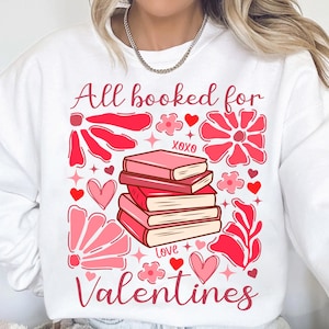 May include: White sweatshirt with a pink and red graphic design featuring a stack of books, flowers, hearts, and the text "All booked for Valentines".