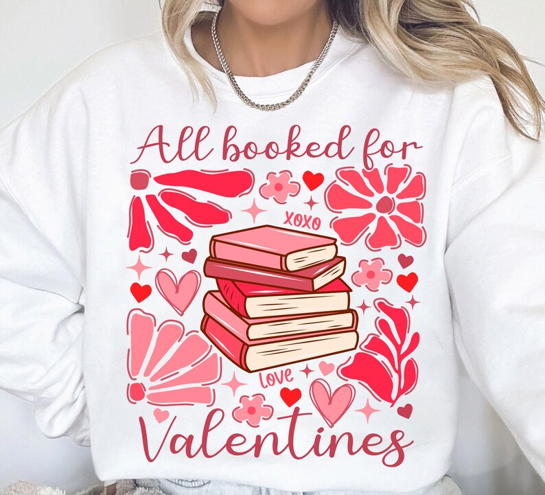 May include: White sweatshirt with a pink and red graphic design featuring a stack of books, flowers, hearts, and the text "All booked for Valentines".