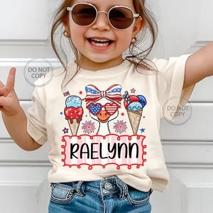 May include: A cream-colored t-shirt featuring a patriotic design with an ice cream cone, a bow, and sunglasses. The shirt has the name "Raelynn" in a red and white frame. The design incorporates red, white, and blue colors.