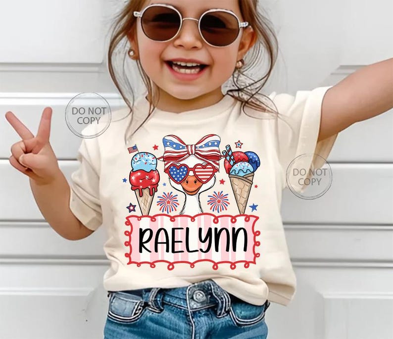 May include: A cream-colored t-shirt featuring a patriotic design with an ice cream cone, a bow, and sunglasses. The shirt has the name "Raelynn" in a red and white frame. The design incorporates red, white, and blue colors.