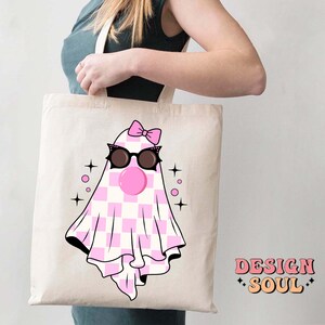 May include: A white tote bag with a pink and white checkered ghost wearing sunglasses and a pink bow. The ghost has a pink bubble coming out of its mouth. The text "DESIGN SOUL" is printed on the bottom right corner of the bag.
