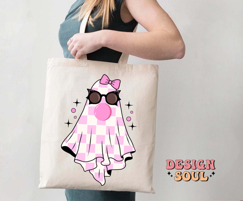 May include: A white tote bag with a pink and white checkered ghost wearing sunglasses and a pink bow. The ghost has a pink bubble coming out of its mouth. The text "DESIGN SOUL" is printed on the bottom right corner of the bag.