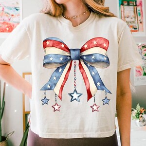 May include: A white t-shirt with a red, white, and blue bow with stars printed on it. The bow has a chain with stars hanging from it.