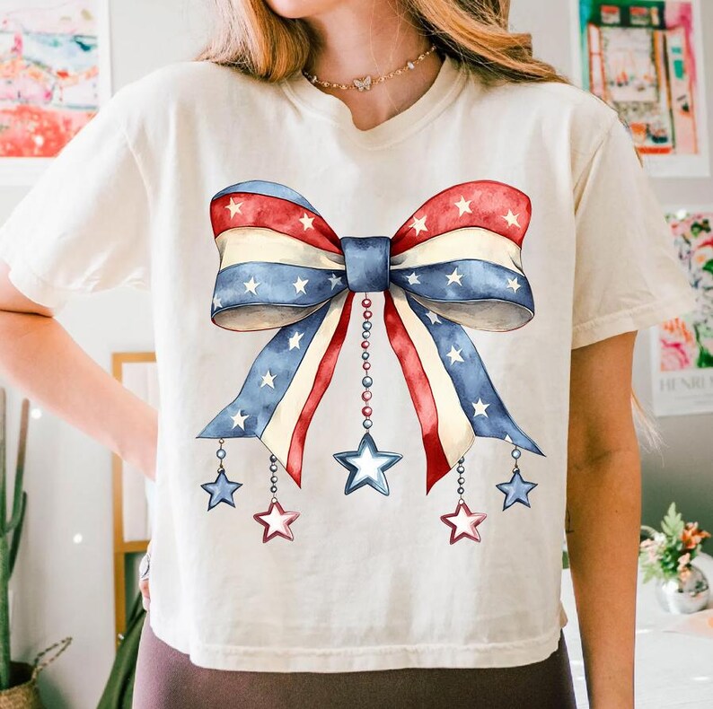 May include: A white t-shirt with a red, white, and blue bow with stars printed on it. The bow has a chain with stars hanging from it.