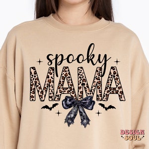 May include: Tan sweatshirt with the text "spooky mama" in black letters with a leopard print pattern. The letters are outlined in black. A black bow with white lace is below the text. Two black bats are on either side of the bow. The text "DESIGN SOUL" is in the bottom right corner.