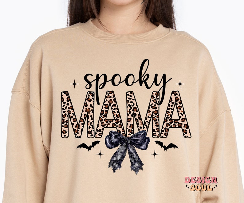 May include: Tan sweatshirt with the text "spooky mama" in black letters with a leopard print pattern. The letters are outlined in black. A black bow with white lace is below the text. Two black bats are on either side of the bow. The text "DESIGN SOUL" is in the bottom right corner.
