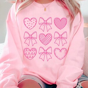 May include: Pink sweatshirt with a graphic design of nine pink hearts and bows. The hearts are outlined in pink and have different patterns inside.