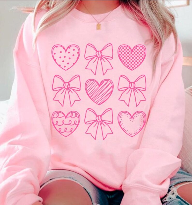May include: Pink sweatshirt with a graphic design of nine pink hearts and bows. The hearts are outlined in pink and have different patterns inside.