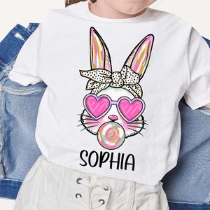 May include: White t-shirt with a colorful bunny wearing pink heart-shaped sunglasses and blowing a pink and green swirl lollipop. The bunny has a black and white polka dot headband. The text "Sophia" is printed below the bunny.