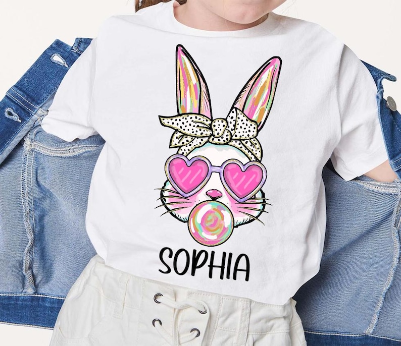 May include: White t-shirt with a colorful bunny wearing pink heart-shaped sunglasses and blowing a pink and green swirl lollipop. The bunny has a black and white polka dot headband. The text "Sophia" is printed below the bunny.