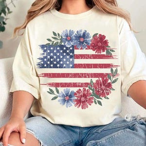 Puede incluir: Camiseta color crema con un dise&ntilde;o desgastado de la bandera estadounidense y detalles florales rojos y azules. La bandera est&aacute; rodeada por una corona de flores en tonos rojos, azules y verdes.