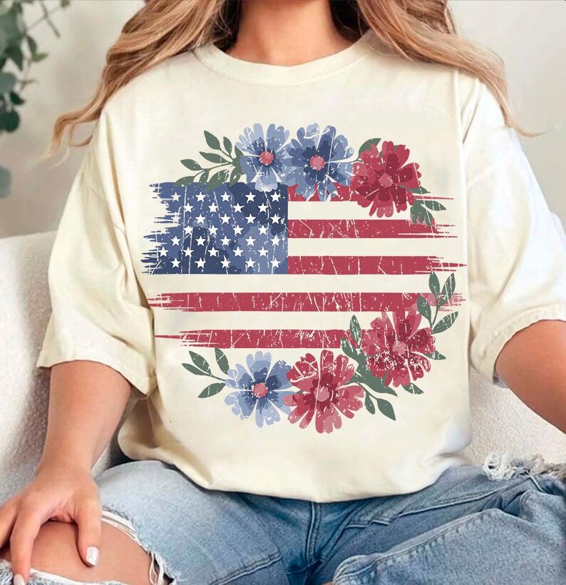 Puede incluir: Camiseta color crema con un dise&ntilde;o desgastado de la bandera estadounidense y detalles florales rojos y azules. La bandera est&aacute; rodeada por una corona de flores en tonos rojos, azules y verdes.