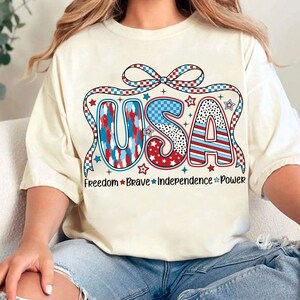 Peut inclure: Un t-shirt blanc avec un graphique USA rouge, blanc et bleu. Le graphique pr&eacute;sente un n&oelig;ud et des &eacute;toiles. Le texte "Freedom Brave Independence Power" est imprim&eacute; sous le graphique.