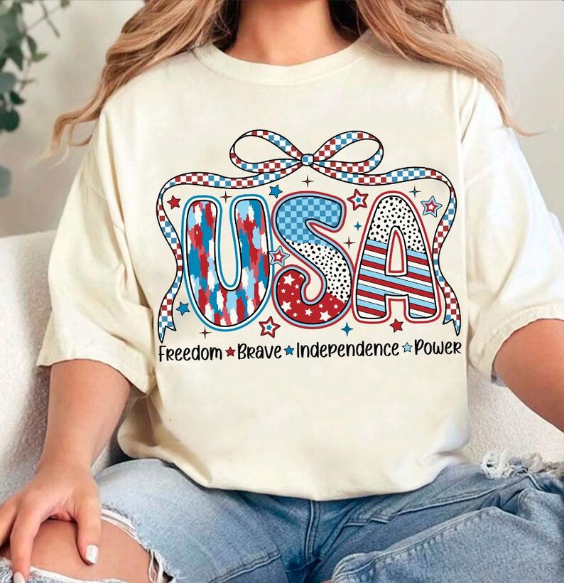 Peut inclure: Un t-shirt blanc avec un graphique USA rouge, blanc et bleu. Le graphique pr&eacute;sente un n&oelig;ud et des &eacute;toiles. Le texte "Freedom Brave Independence Power" est imprim&eacute; sous le graphique.