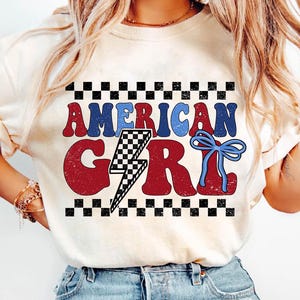 May include: A white t-shirt with a red, white, and blue graphic that says "American Girl" with a lightning bolt and a blue bow. The graphic is surrounded by a black and white checkered border.