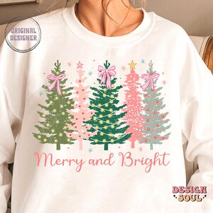 May include: White sweatshirt with a Christmas tree design and the text "Merry and Bright". The trees are green, pink, and blue with pink bows and white lights.