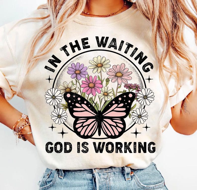 May include: A white t-shirt with a floral design and a butterfly. The text "In the waiting, God is working" is printed on the shirt.