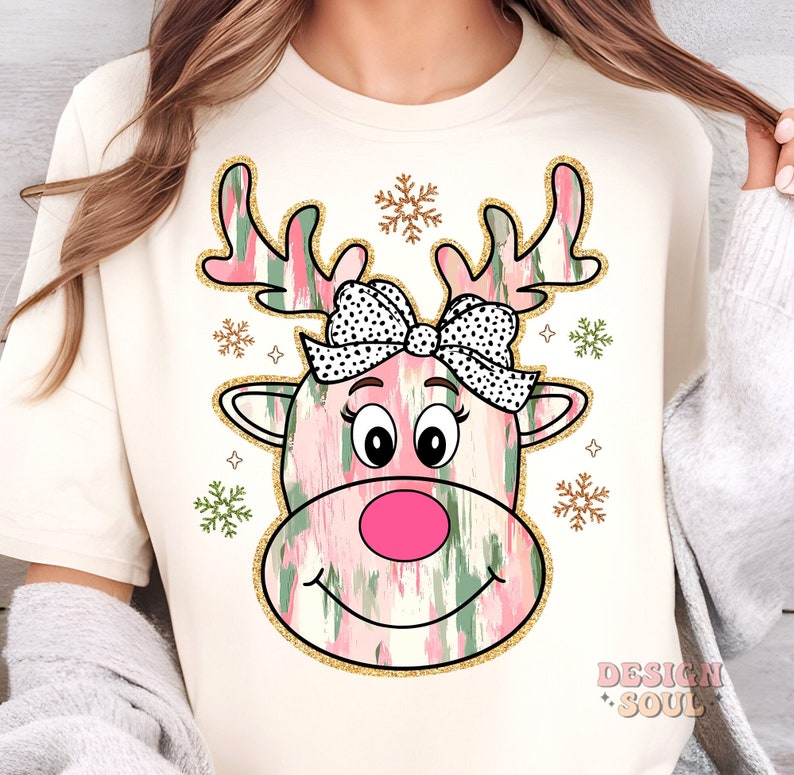 May include: A white t-shirt with a pink and green reindeer design. The reindeer has a black and white polka dot bow and a pink nose. The design is surrounded by gold glitter snowflakes.