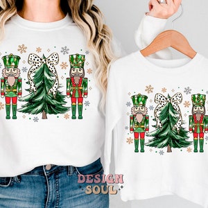 May include: White sweatshirts with a festive Christmas design. The design includes two nutcrackers, a Christmas tree, and a polka-dot bow. The sweatshirts are decorated with snowflakes, ideal for the holiday season.