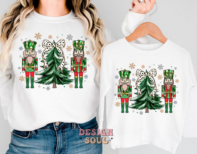 May include: White sweatshirts with a festive Christmas design. The design includes two nutcrackers, a Christmas tree, and a polka-dot bow. The sweatshirts are decorated with snowflakes, ideal for the holiday season.