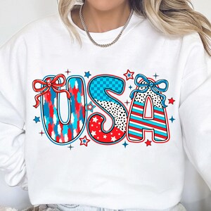 May include: White sweatshirt with a red, white, and blue USA graphic. The letters are decorated with stars, stripes, and polka dots.