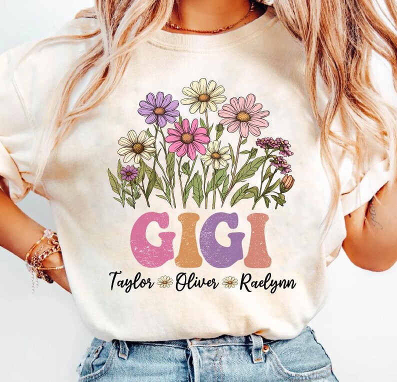 May include: Cream-colored t-shirt featuring a floral design with daisies and other flowers in shades of pink, purple, and yellow. The word "GIGI" is printed in a retro font, with names below. A personalized gift.
