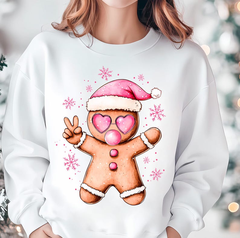 May include: White sweatshirt with a gingerbread man wearing a pink Santa hat and pink heart-shaped sunglasses. The gingerbread man is holding up two fingers in a peace sign. The background is white with pink snowflakes.