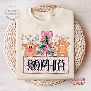 Op de afbeelding: Een beige sweatshirt met een roze en wit kerstontwerp. Het ontwerp bevat een rendier, een peperkoekman en een kerstboom. De tekst "Sophia" is geschreven in zwarte letters op een roze en witte luipaardprint achtergrond.