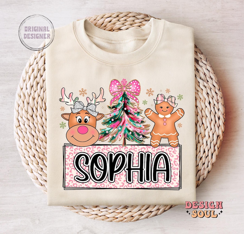 Op de afbeelding: Een beige sweatshirt met een roze en wit kerstontwerp. Het ontwerp bevat een rendier, een peperkoekman en een kerstboom. De tekst "Sophia" is geschreven in zwarte letters op een roze en witte luipaardprint achtergrond.
