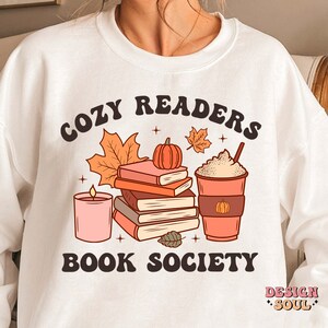 May include: White sweatshirt with a graphic design of a stack of books, a candle, a cup of coffee, and fall leaves. The text "Cozy Readers Book Society" is printed on the sweatshirt.