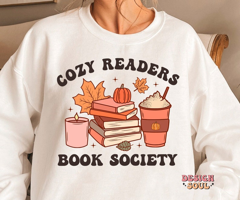 May include: White sweatshirt with a graphic design of a stack of books, a candle, a cup of coffee, and fall leaves. The text "Cozy Readers Book Society" is printed on the sweatshirt.