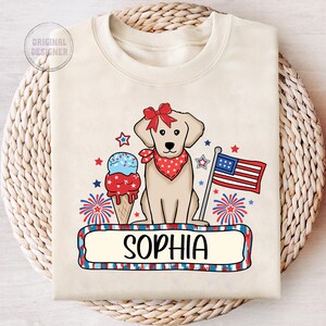 Pu&ograve; includere: Una maglietta beige con un designo patriottico rosso, bianco e blu che presenta un cane, un cono gelato, fuochi d'artificio e una bandiera. Il testo "Sophia" &egrave; stampato su uno striscione sotto il designo.