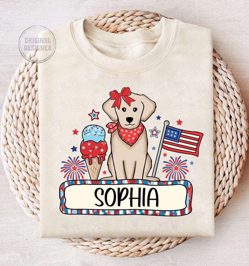 Pu&ograve; includere: Una maglietta beige con un designo patriottico rosso, bianco e blu che presenta un cane, un cono gelato, fuochi d'artificio e una bandiera. Il testo "Sophia" &egrave; stampato su uno striscione sotto il designo.