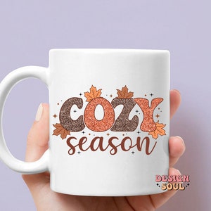 May include: White ceramic mug with the words "COZY season" in brown and orange glitter lettering, decorated with fall leaves. The mug is held by a hand.