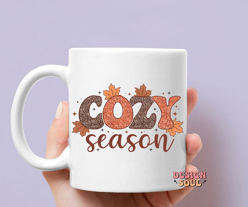 May include: White ceramic mug with the words "COZY season" in brown and orange glitter lettering, decorated with fall leaves. The mug is held by a hand.