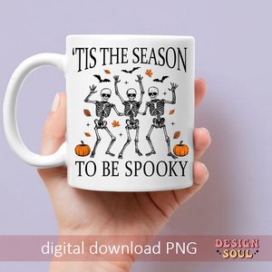 May include: White ceramic mug with a black graphic of three skeletons dancing with the text 'Tis the season to be spooky' and two orange pumpkins.