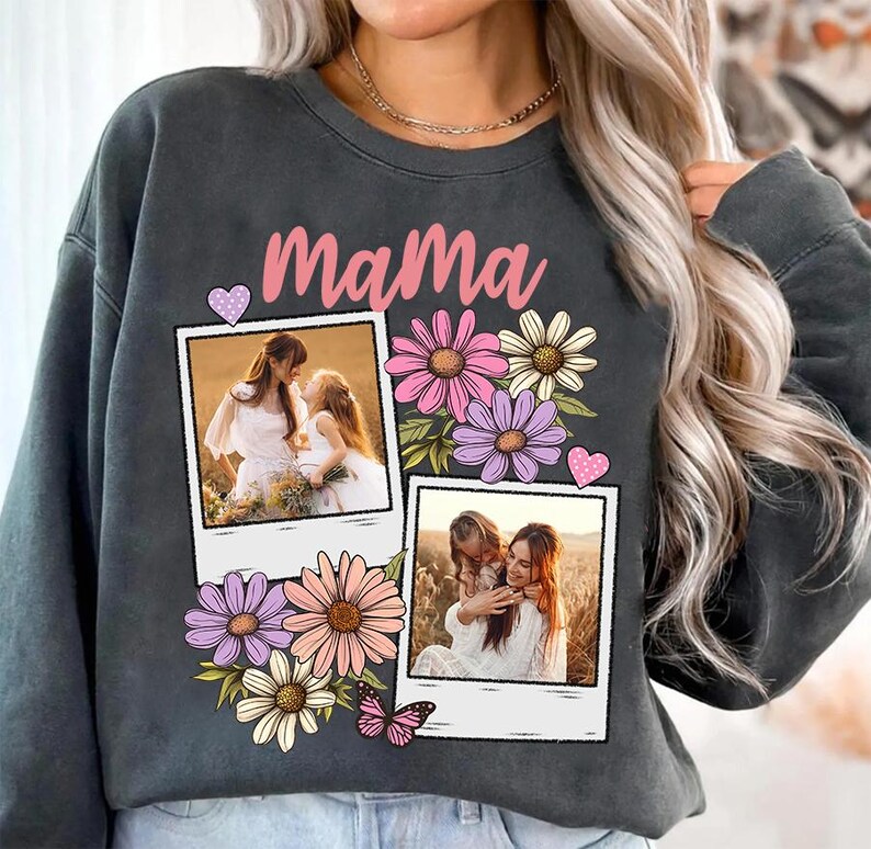 May include: Dark gray sweatshirt with the word "Mama" in pink script. The sweatshirt features two polaroid-style photos, floral accents, and a butterfly graphic. The photos show a mother and child.