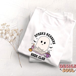 May include: White t-shirt with a graphic of a ghost reading a book with a candle and a pumpkin. The text "Spooky Readers Book Club" is in a circle around the ghost.