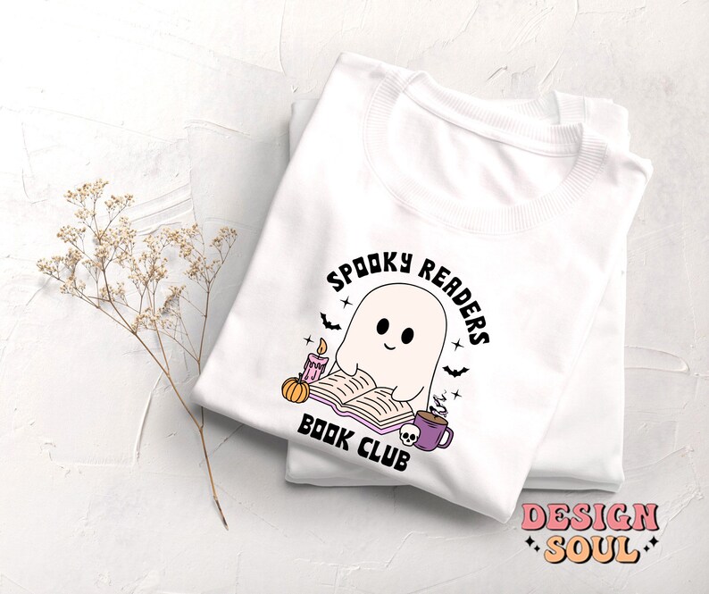 May include: White t-shirt with a graphic of a ghost reading a book with a candle and a pumpkin. The text "Spooky Readers Book Club" is in a circle around the ghost.