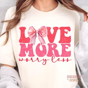 May include: A white t-shirt with a pink and red graphic that says "Love More Worry Less". A pink watercolor bow is above the word "Love".