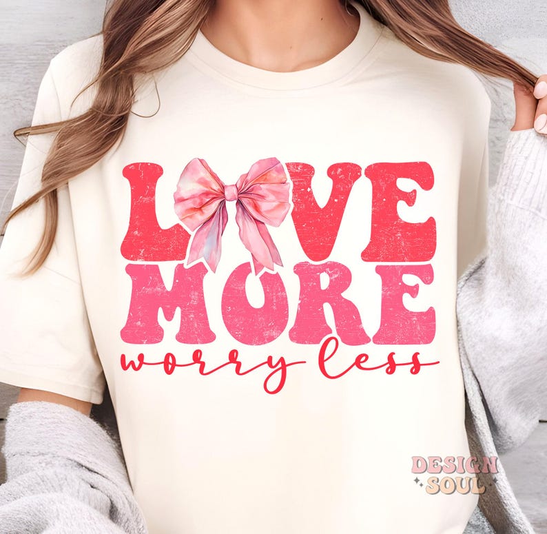 May include: A white t-shirt with a pink and red graphic that says "Love More Worry Less". A pink watercolor bow is above the word "Love".