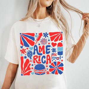 以下が含まれることがあります： 星、花火、ハート、その他の愛国的なイメージに囲まれた「AMERICA」という言葉が描かれた、赤、白、青のグラフィックデザインが施された白いTシャツです。
