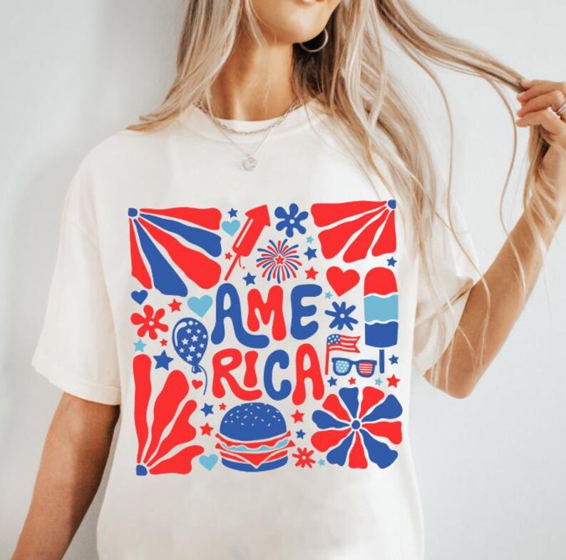 以下が含まれることがあります： 星、花火、ハート、その他の愛国的なイメージに囲まれた「AMERICA」という言葉が描かれた、赤、白、青のグラフィックデザインが施された白いTシャツです。