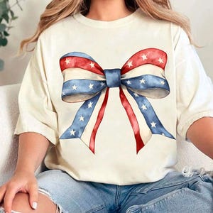 K&ouml;nnte beinhalten: Ein wei&szlig;es T-Shirt mit einer rot-wei&szlig;-blauen Schleife mit Sternen darauf gedruckt. Die Schleife ist im Aquarell-Stil gehalten.