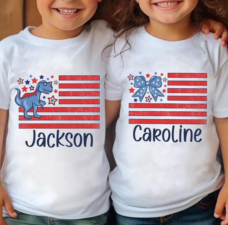 May include: Two white t-shirts with an American flag design. One shirt features a blue dinosaur with sunglasses and the name "Jackson". The other shirt features a blue bow and the name "Caroline".