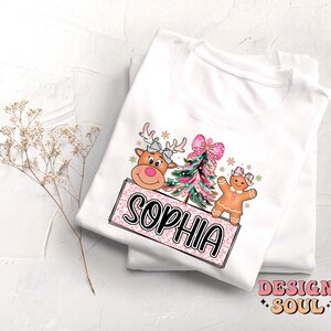 Op de afbeelding: Wit T-shirt met een roze en wit ontwerp met een rendier, een kerstboom en een peperkoekman. Het ontwerp bevat ook de naam "Sophia".