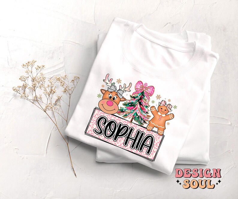 Op de afbeelding: Wit T-shirt met een roze en wit ontwerp met een rendier, een kerstboom en een peperkoekman. Het ontwerp bevat ook de naam "Sophia".