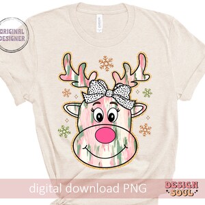 May include: A digital download PNG image of a pink and green reindeer with a pink bow and snowflakes. The reindeer has a pink nose and black eyes. The background is a beige t-shirt.
