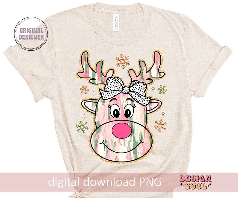 May include: A digital download PNG image of a pink and green reindeer with a pink bow and snowflakes. The reindeer has a pink nose and black eyes. The background is a beige t-shirt.
