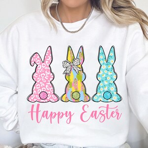 May include: White sweatshirt with a pink, blue, and yellow bunny rabbit design. The bunnies are wearing bows and have different patterns. The text "Happy Easter" is printed in pink below the bunnies.